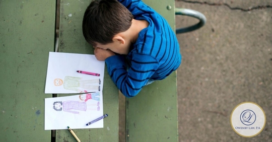 Little boy with his head down on a table with crayons and a drawing next to himof mom, dad, and child cut in half.